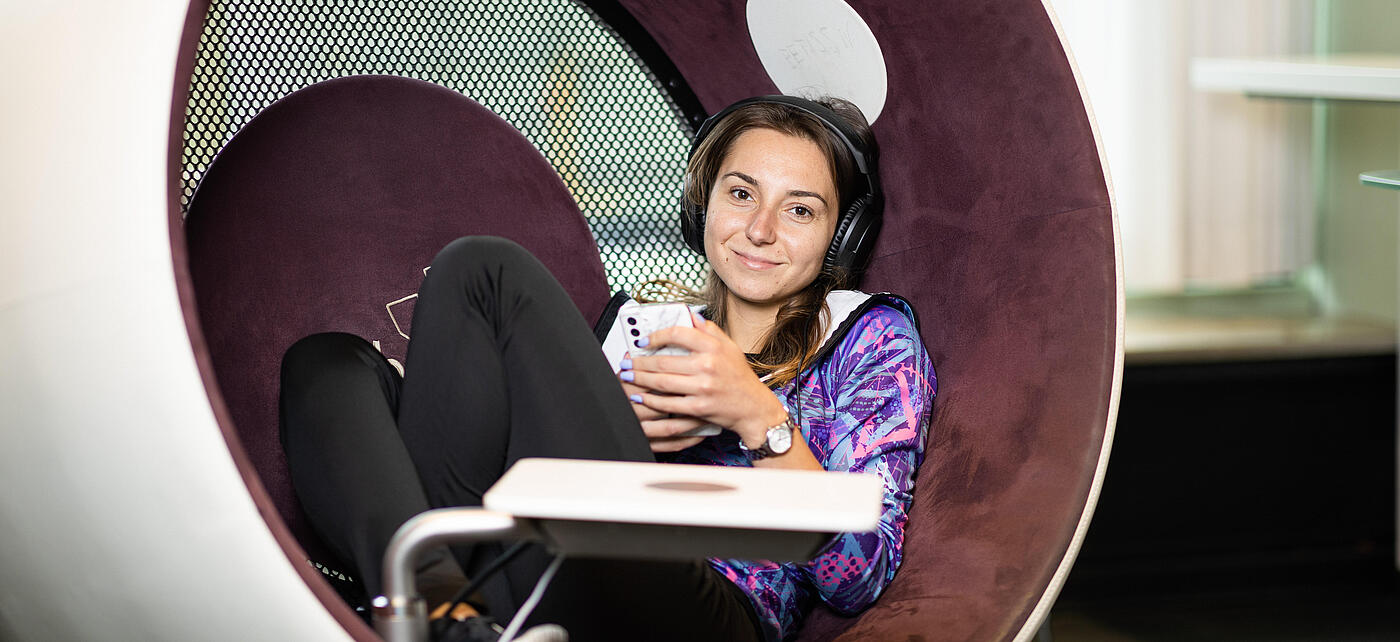 Young woman with headphones and smartphone sits in sonic chair and smiles at camera