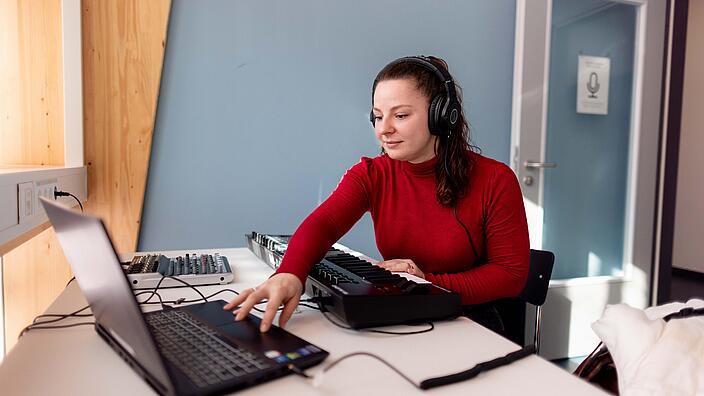 Frau sitzt mit Kopfhörer an Schreibtisch und arbeitet mit Laptop und Piano