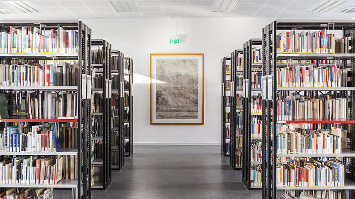 Rechts und links Regale voller Bücher, an Wand am Ende des Raumes gerahmte historische Karte