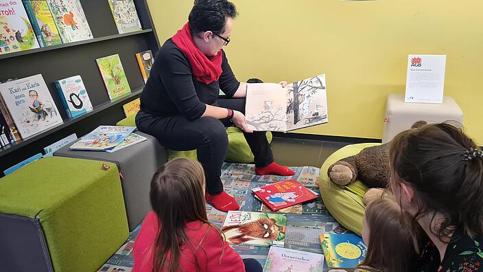 Auf einem Teppich liegen verschiedene Bilderbücher, eine Dame hält eins in der Hand und schaut es sich mit Kindern an