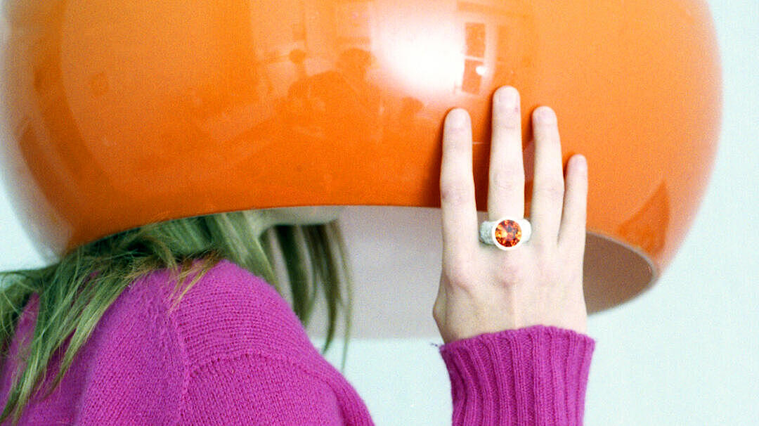 Eine weibliche Person ist unter einem orangenen Lampenschrim versteckt. Ihr lilaner Pullover und ihre Hand, die den Schirm festhält dominieren das Foto.  