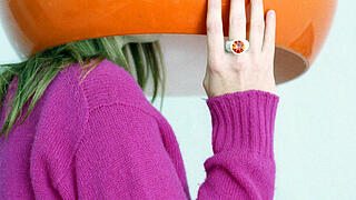 Eine weibliche Person ist unter einem orangenen Lampenschrim versteckt. Ihr lilaner Pullover und ihre Hand, die den Schirm festhält dominieren das Foto.