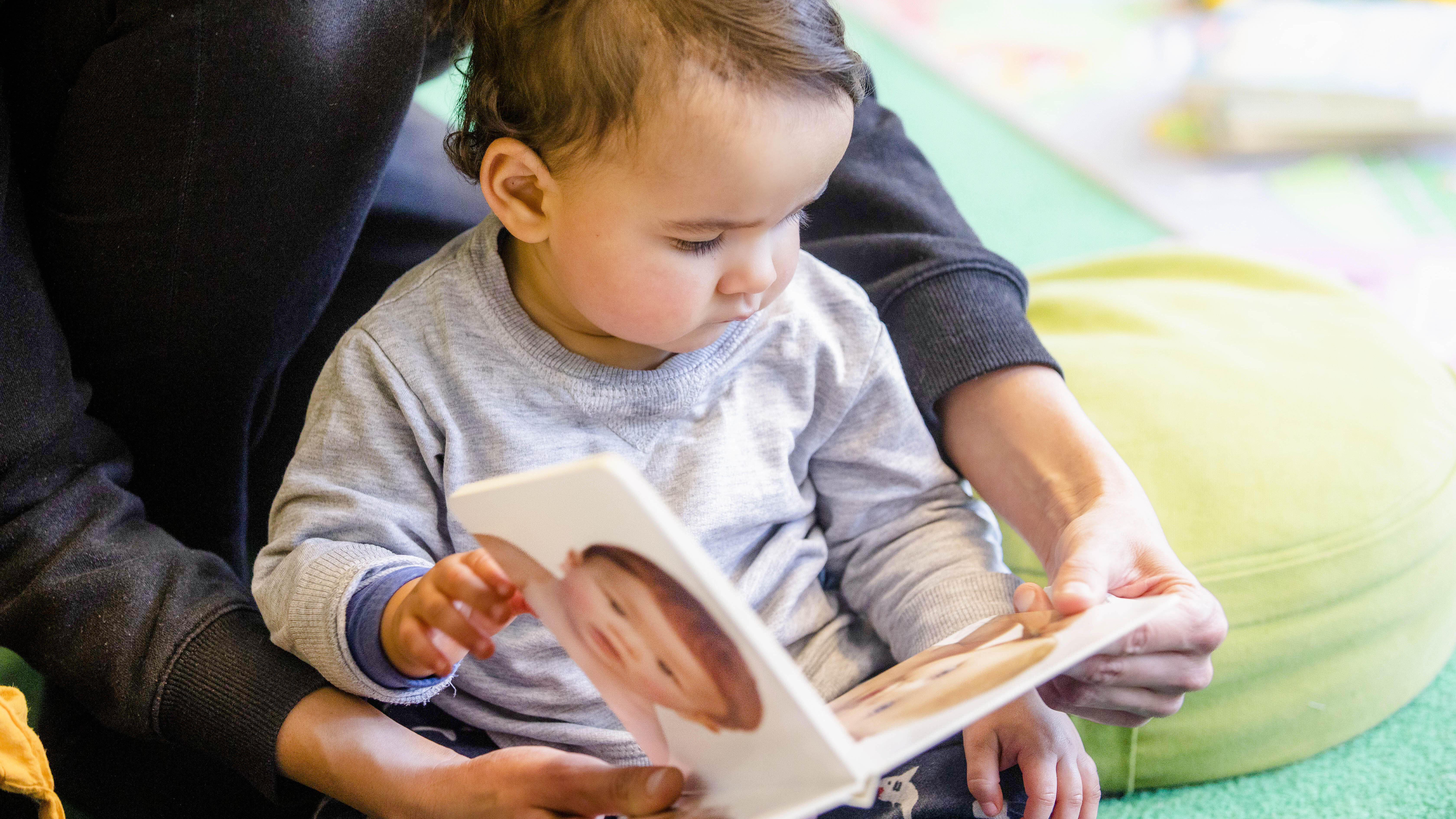 Kleinkind sitzt auf Boden in Bibliothek und betrachtet ein Babybuch, das Erwachsener festhält
