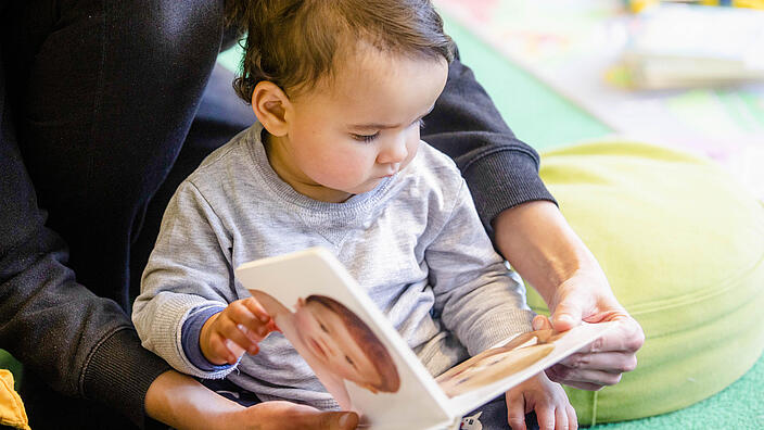 Kleinkind sitzt auf Boden in Bibliothek und betrachtet ein Babybuch, das Erwachsener festhält