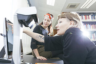 Zwei Frauen sitzen in der Bibliothek und schauen gemeinsam auf einen PC
