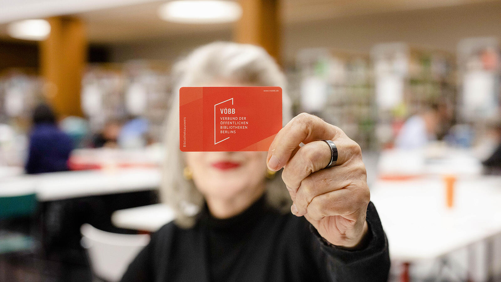 Older lady holding red library card to camera covering her face