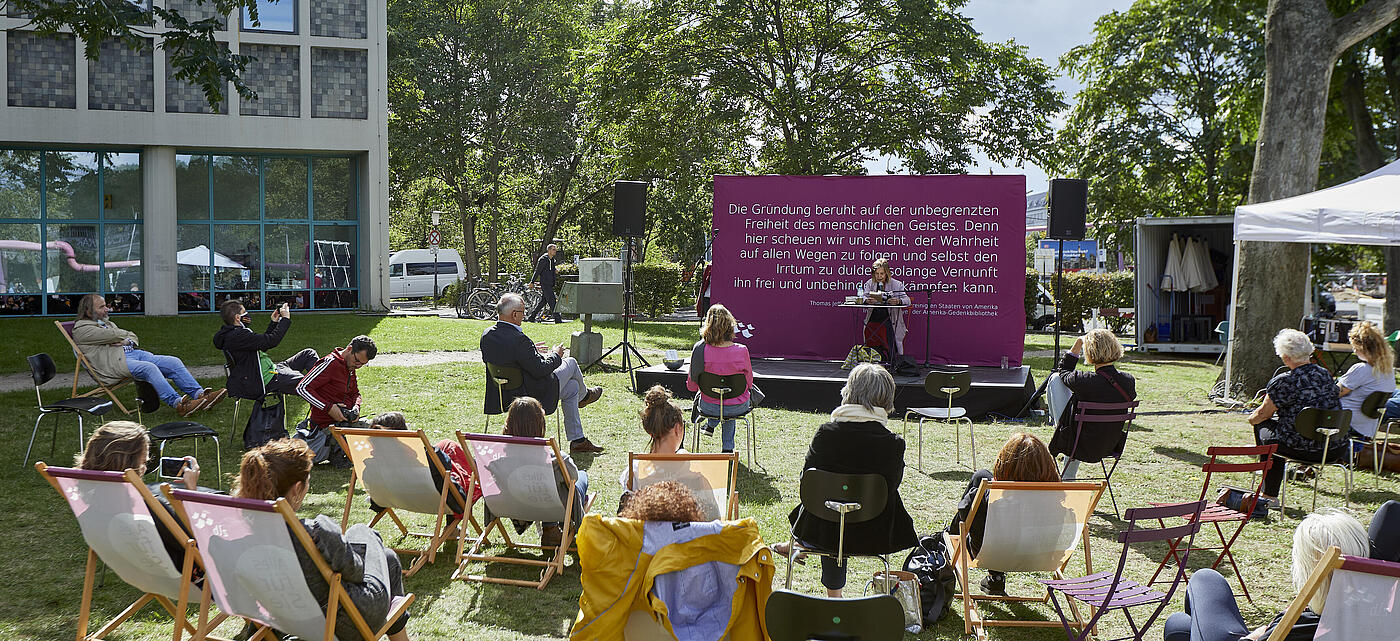 People sitting in deck chairs on lawn in front of small stage with speaker at microphone