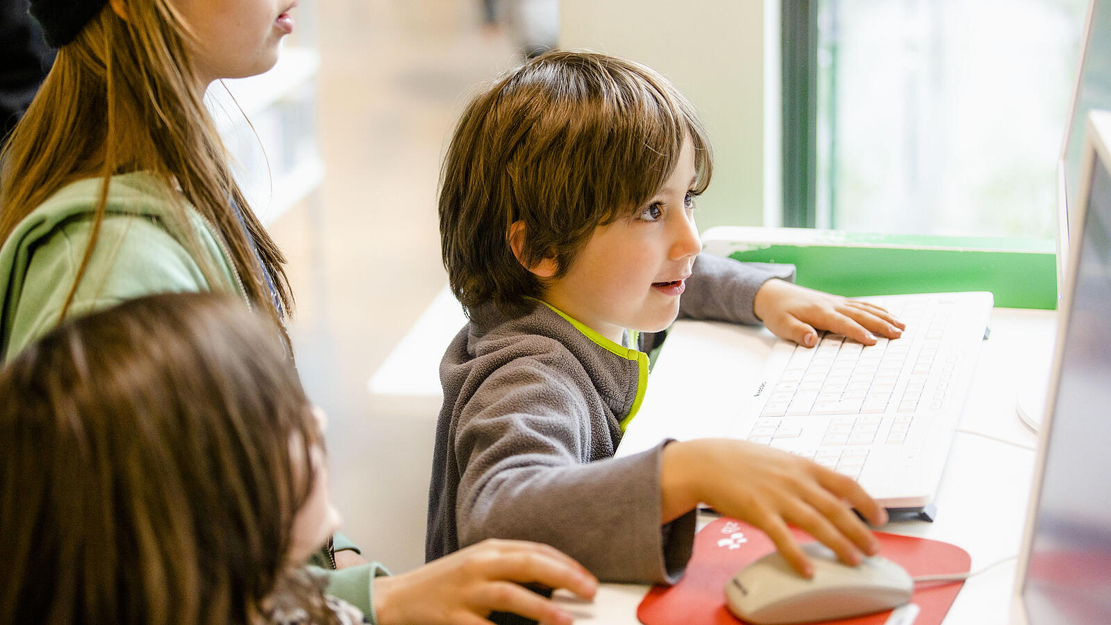 Kleines Kind steht neben anderen Kindern vor Schreibtisch und blickt gespannt auf Computerbildschirm