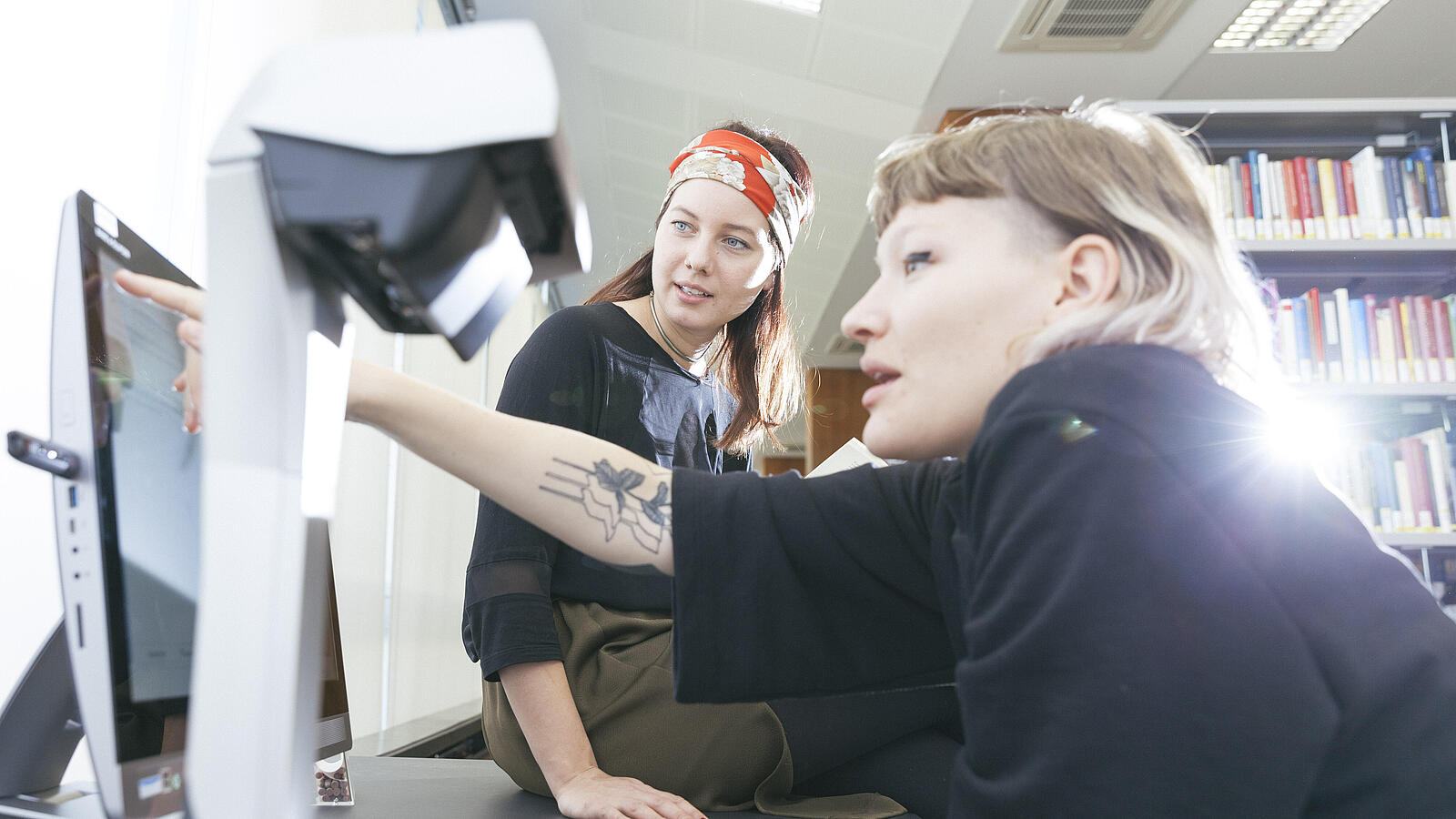 Eine Frau zeigt einer anderen Frau eine bestimmte Stelle auf Computerbildschirm in Bibliothek