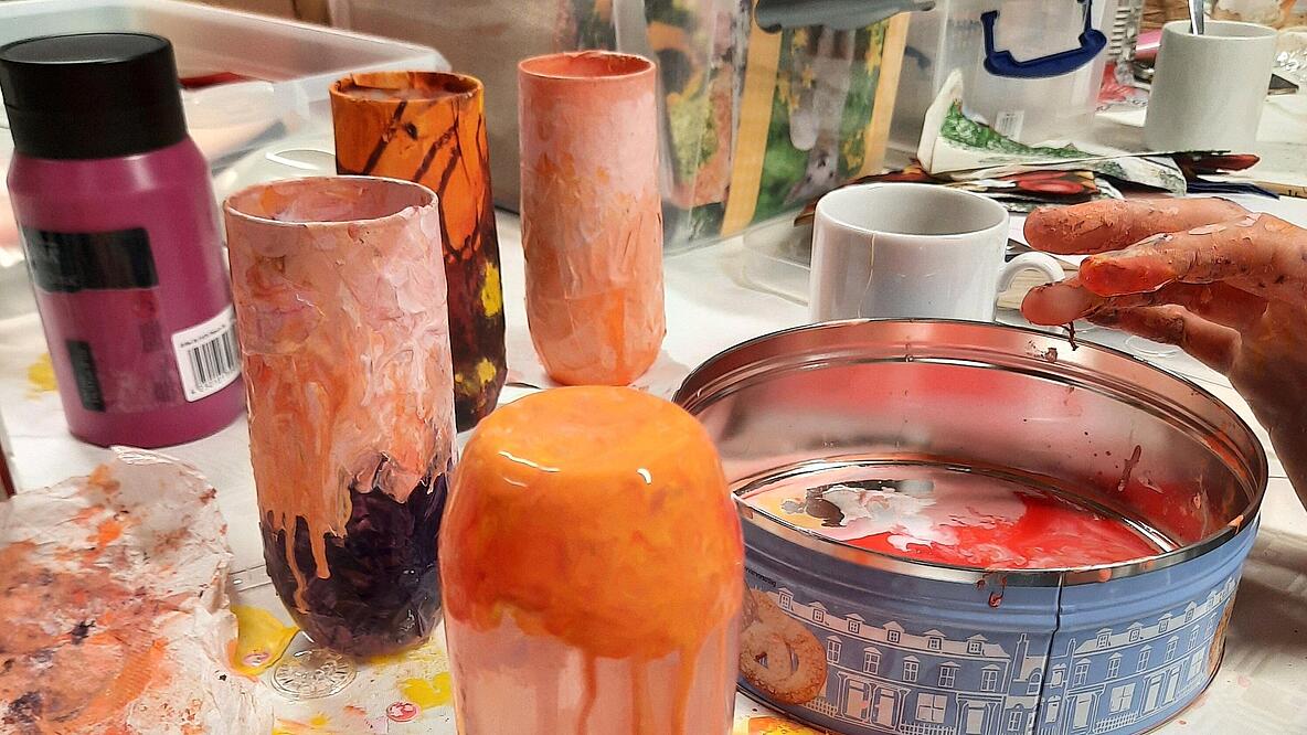 Bildausschnitt Hände bemalen mit orangener Farbe ein Glas. Im Hintergrund stehen fertig gestaltete Gläser.