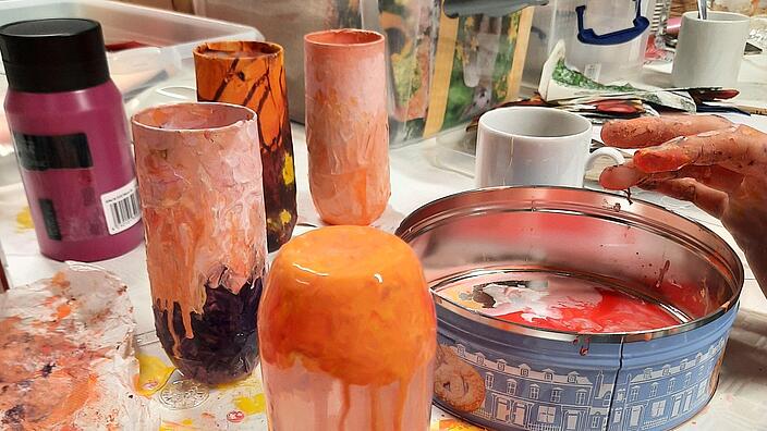 Bildausschnitt Hände bemalen mit orangener Farbe ein Glas. Im Hintergrund stehen fertig gestaltete Gläser.