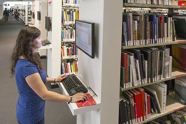 Frau steht in der Lesehalle der AGB und recherchiert an PC Katalog