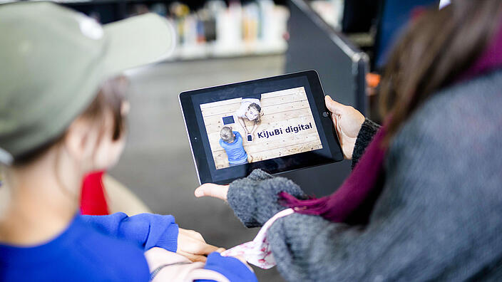 Junge Frau und Mädchen schauen auf ein Tablet, das ein Bild mit Aufschrift "KiJuBi digital" anzeigt