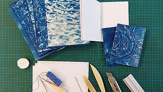 mehrere selbstgebundene Blaudrucke liegen zusammen mit Buchbindewerkzeugen wie beispielsweise Pfalzbeilen, Nadeln und einem Lineal auf einer Schneidematte ausgebreitet
