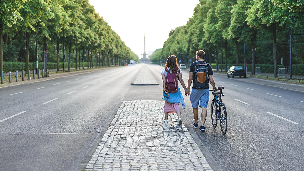 Auf einer breiten Allee gesäumt von grünen Bäumen laufen ein Mann und eine Frau händchenhaltend auf der Fahrbahn. Die Straße ist auto- und menschenleer. In der Ferne lässt sich die Siegessäule erkennen. 