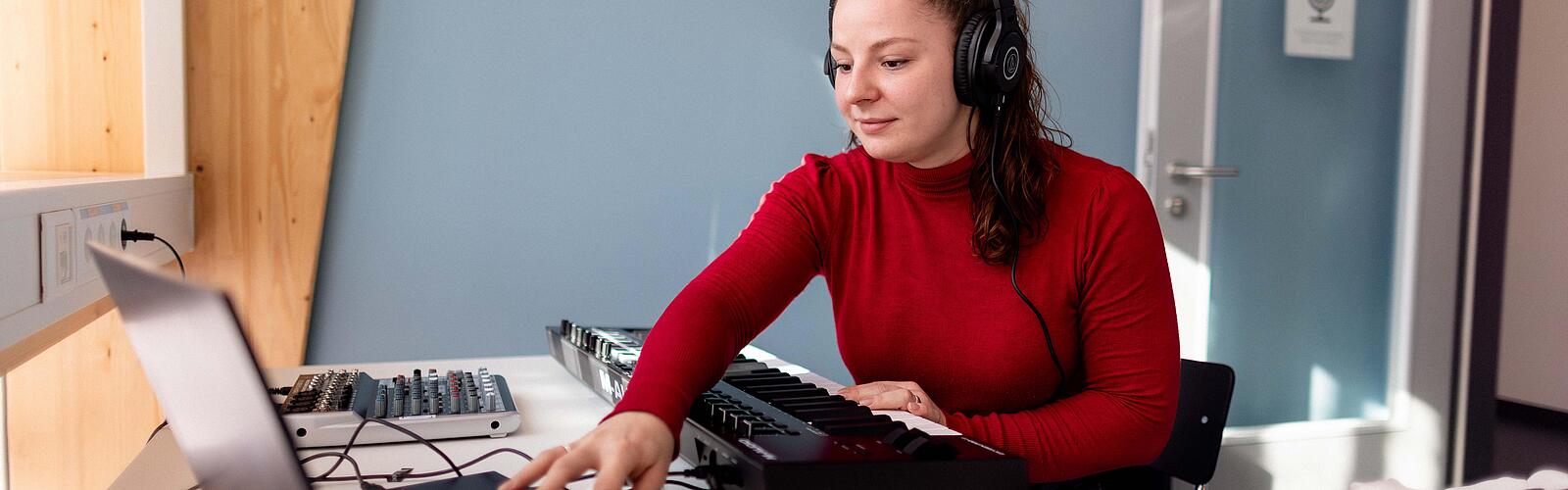 Frau sitzt mit Kopfhörer an Schreibtisch und arbeitet mit Laptop und Piano