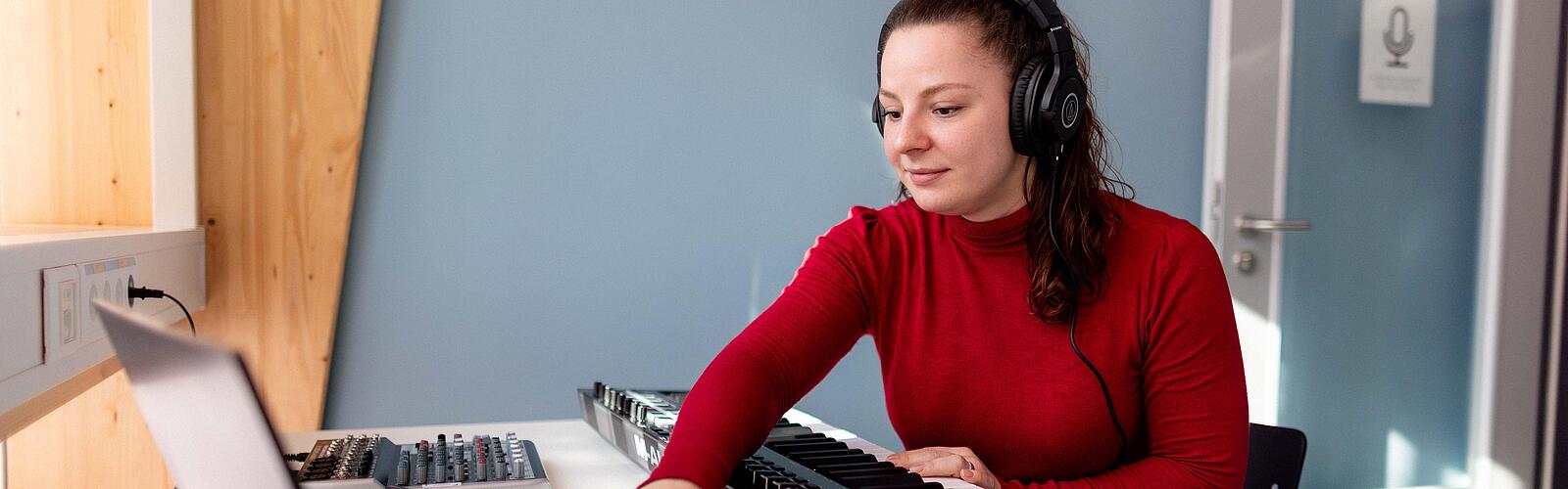 Frau sitzt mit Kopfhörer an Schreibtisch und arbeitet mit Laptop und Piano