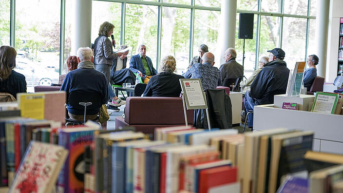 Auf einem Foto sieht man eine Veranstaltung im Salon der Amerika-Gedenkbibliothek. Im Vordergrund steht ein Tisch vollter bunter Bücher. Im Hintergrund sind Menschen zu erkennen. Vor einem Podium sitzt ein dutzend Menschen und hört der Moderatorin zu. 
