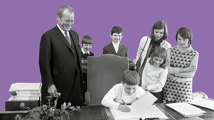 Bundeskanzler a.D. Willy Brandt steht an einem Schreibtisch. An dem Schreibtisch sitzt ein Kind und unterzeichnet lachend ein Dokument. Um den Schreibtisch herum stehen weitere drei lachende Kinder und zwei erwachsene Personen. Alle blicken zu dem Kind, das am Schreibtisch sitzt.