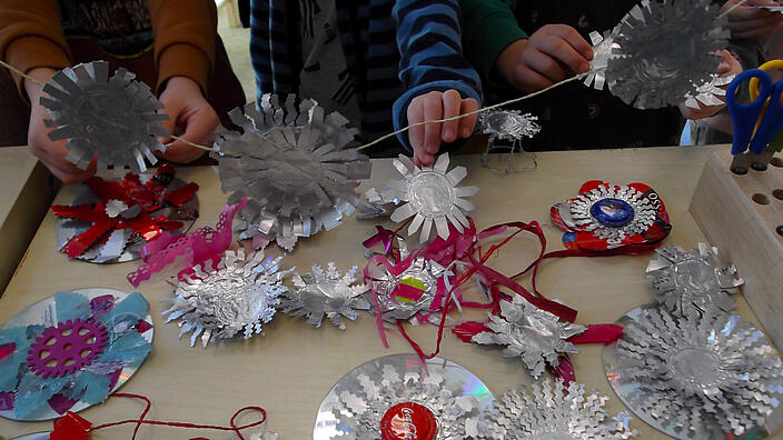 mehrere Kinderhände basteln Objekte aus CD´s, Alufolie, Kronenkorken und anderen recycelten Materialien