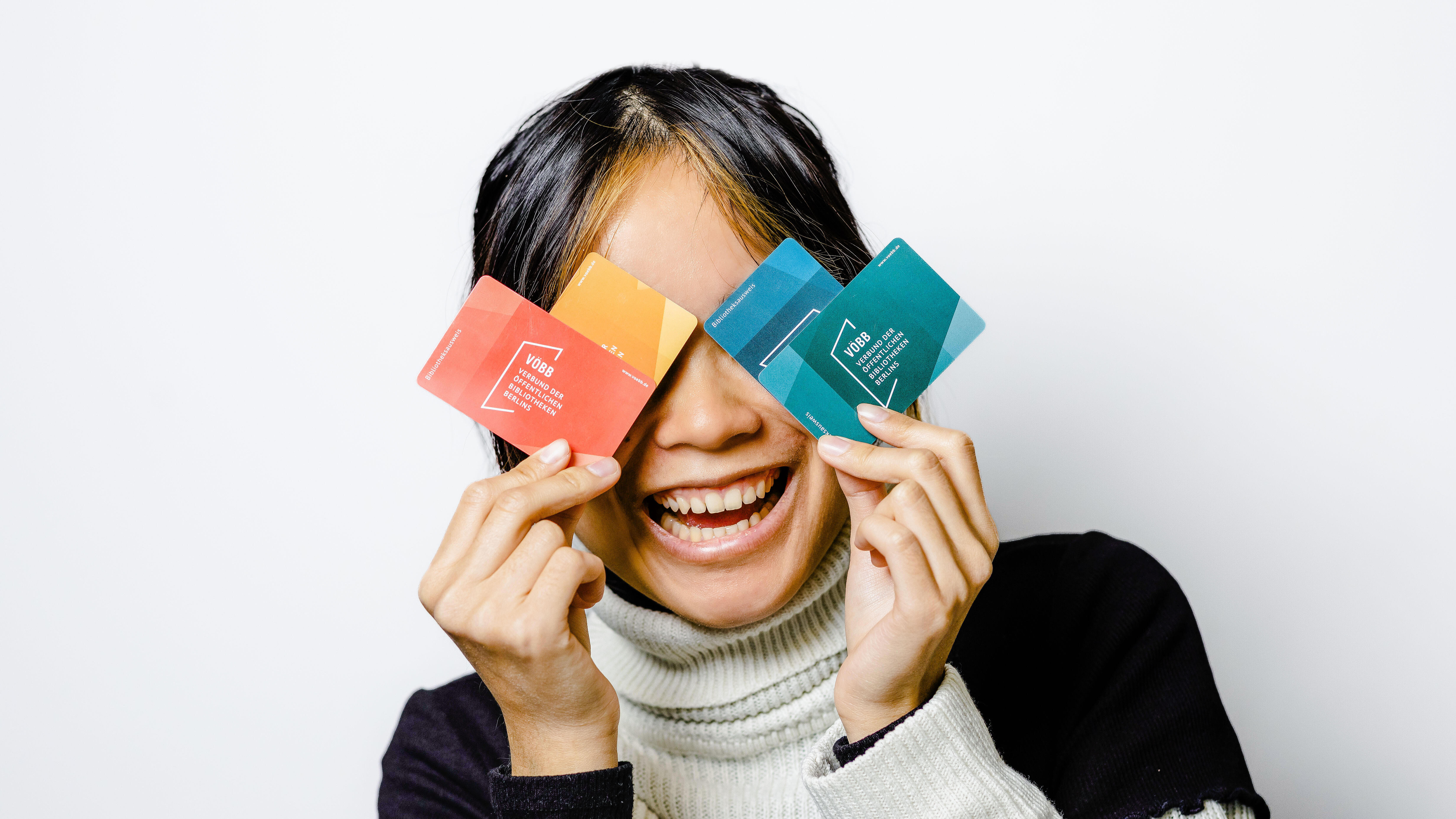 Junge Frau hält lachend vier verschiedenfarbige Varianten des Bibliotheksausweises vor ihre Augen