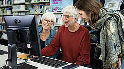 Junge Frau und älteres Ehepaar schauen gemeinsam auf einen Bildschirm und Lachen