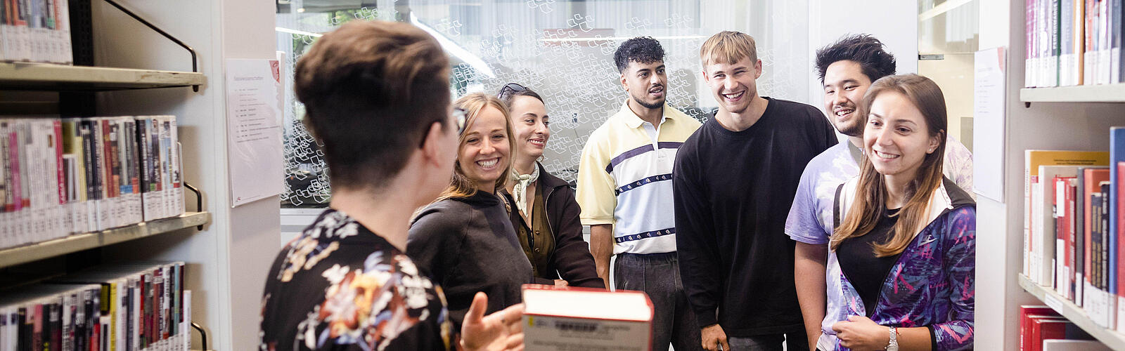 Frau zeigt einer Gruppe junger fröhlicher Menschen ein Buch in der Bibliothek