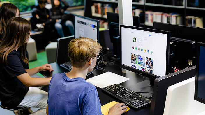Jugendliche sitzen an Schreibtisch vor Computerbildschirmen in Bibliothek