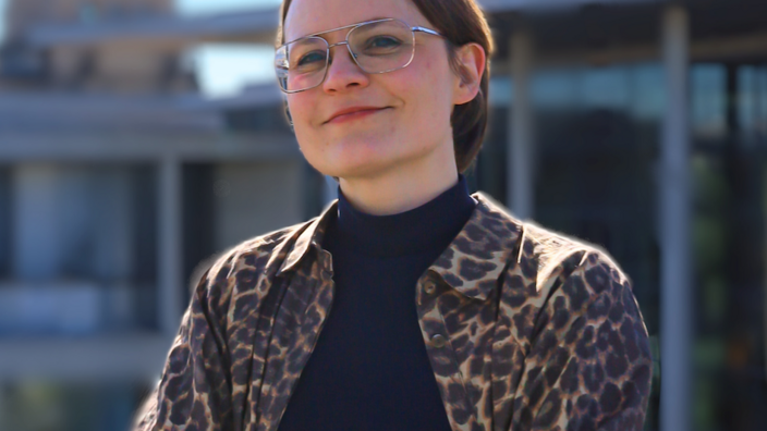 Jo Lücke steht auf einer Terasse und schaut mit verschrenkten Armen in die Kamera. Sie trägt die Haare kurz, eine Brille und eine Jacke im Leo-Print. Im Hintergrund sieht man blauen Himmel und Gebäude. 