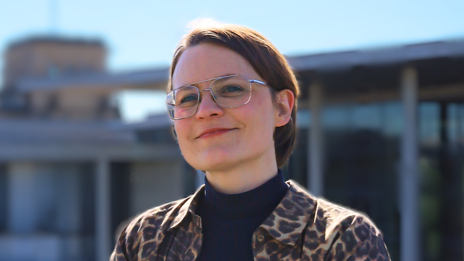 Jo Lücke steht auf einer Terasse und schaut mit verschrenkten Armen in die Kamera. Sie trägt die Haare kurz, eine Brille und eine Jacke im Leo-Print. Im Hintergrund sieht man blauen Himmel und Gebäude. 