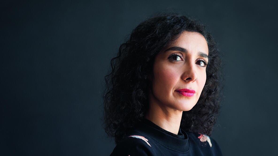 Vor einem grauen Hintergrund steht Asha Hedayati. Sie trägt eine schwarze Bluse und offene schwarze Locken. Sie schaut klug und schräg in die Kamera. 