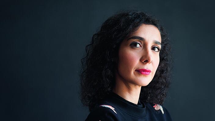 Vor einem grauen Hintergrund steht Asha Hedayati. Sie trägt eine schwarze Bluse und offene schwarze Locken. Sie schaut klug und schräg in die Kamera. 