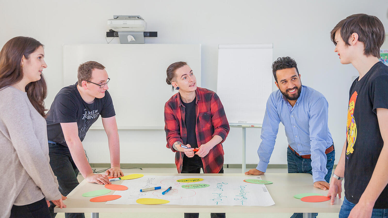 Gruppe Junger Menschen steht in einem Raum um einen Tisch und diskutiert über Workshopinhalte
