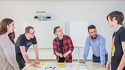 Gruppe Junger Menschen steht in einem Raum um einen Tisch und diskutiert über Workshopinhalte
