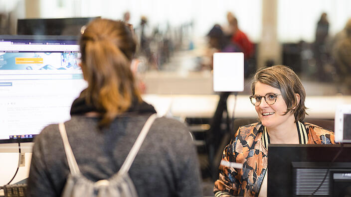 Woman at computer reacts to woman standing in front of consulting counter