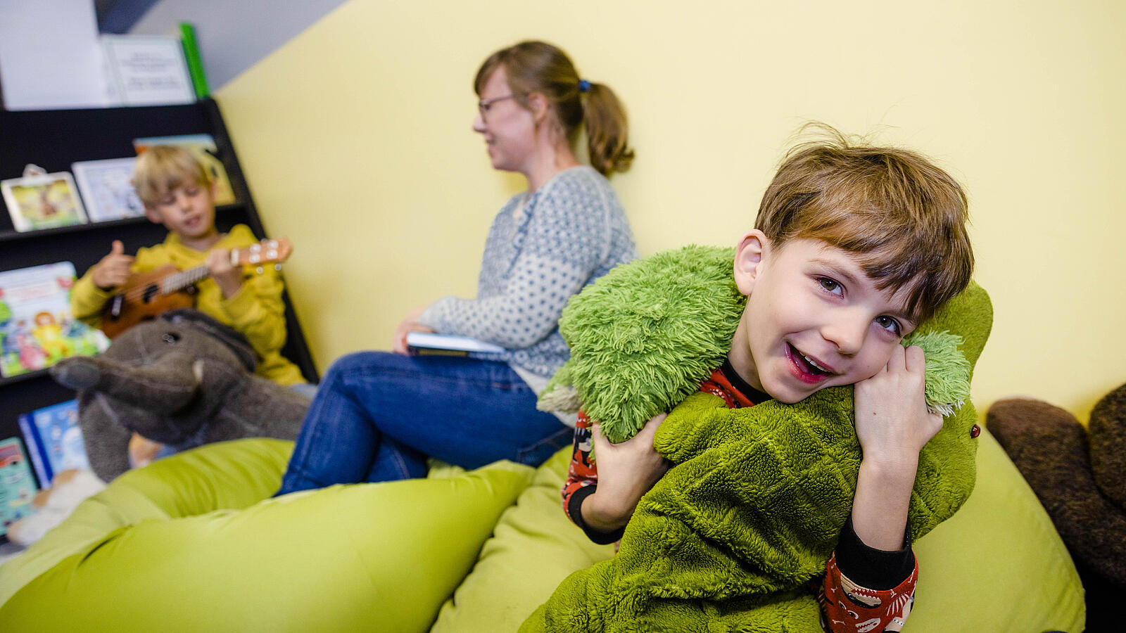 Zwei Jungen spielen mit Kuscheltier und Ukulele mit einer Frau in gemütlicher Ecke in Bibliothek