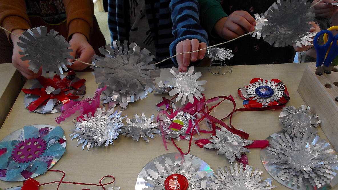 mehrere Kinderhände basteln Objekte aus CD´s, Alufolie, Kronenkorken und anderen recycelten Materialien