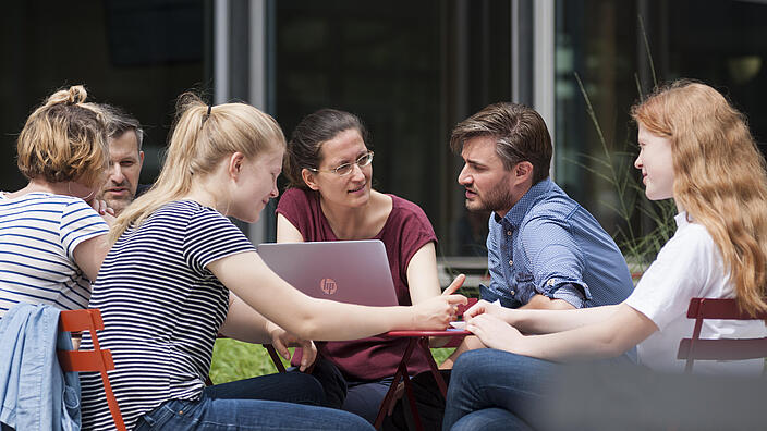 Menschen sitzen im Innenhof gemeinsam an einem Tisch mit Laptop und unterhalten sich