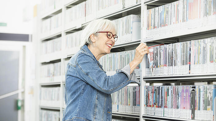 Ältere Frau zieht lächelnd DVD aus Regal in Bibliothek