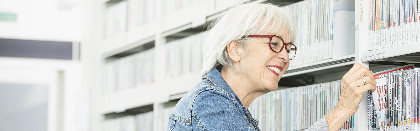 Ältere Frau zieht lächelnd DVD aus Regal in Bibliothek