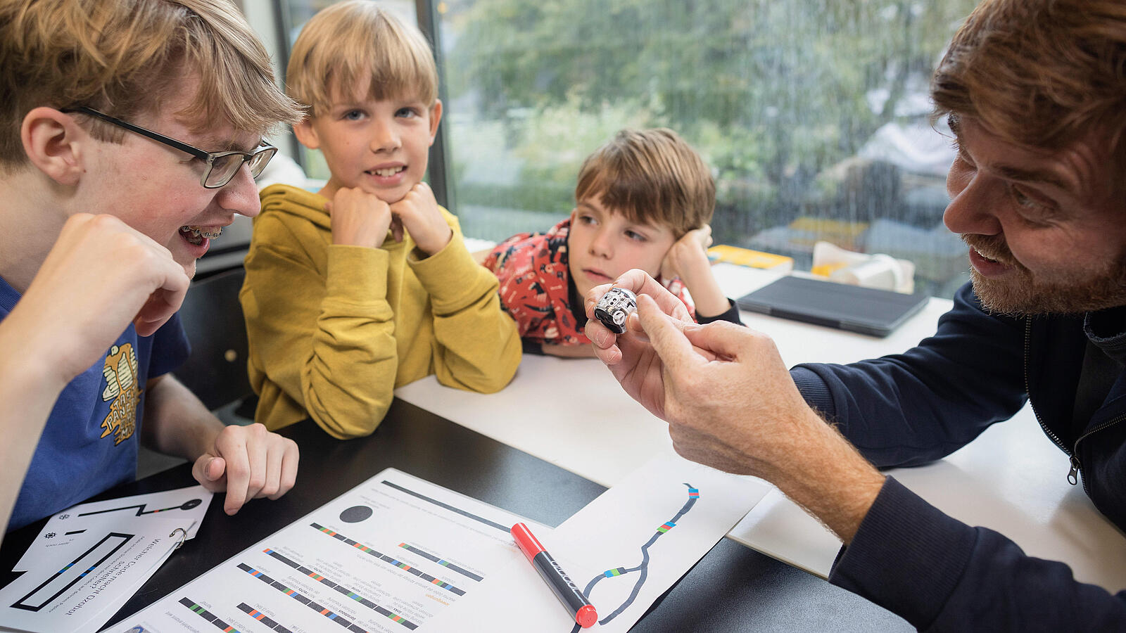 Lernsituation mit 3 Schülern und einem Lehrer, der an einem kleinen Roboter etwas demonstriert