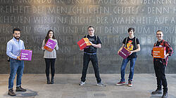 Foto von fünf jungen Personen im Halbkreis aufgestellt im Foyer der AGB mit großen Würfeln mit der Aufschrift "Raum für Neues" in den Händen