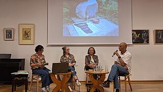 Vier Personen sitzen bei einer Veranstaltung, eine Person spricht mit Mikrofon. Im Hintergrund ist das Herero-Nama-Denkmal auf einer Leinwand zu sehen.