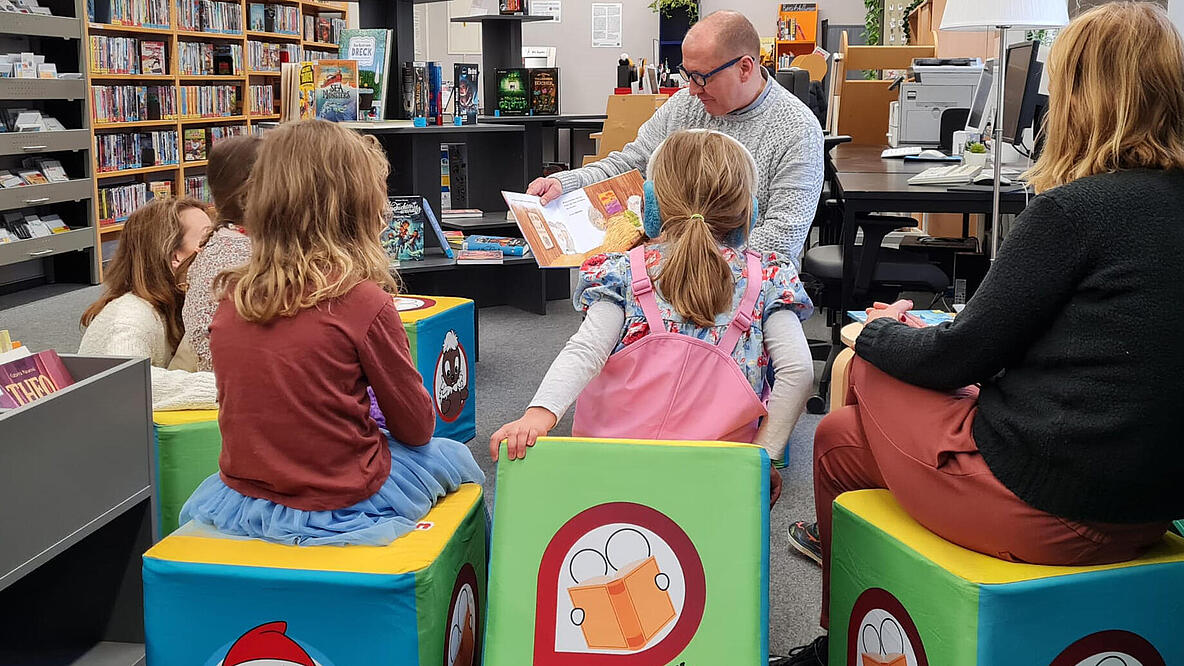 Kinder und Erwachsene sitzen mit dem Rücken zum Betrachter in einer Bibliothek auf Sitzwürfeln und bekommen eine Geschichte von einem Erwachsenen, der vor ihnen sitzt, vorgelesen