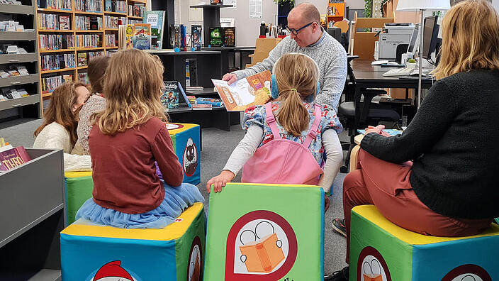 Kinder und Erwachsene sitzen mit dem Rücken zum Betrachter in einer Bibliothek auf Sitzwürfeln und bekommen eine Geschichte von einem Erwachsenen, der vor ihnen sitzt, vorgelesen