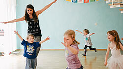vier Kinder tanzen im Raum verstreut in Begleitung einer Frau durch den Raum