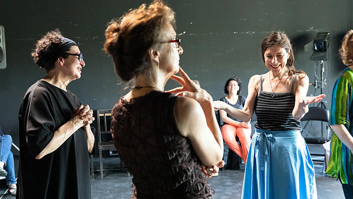 Auf dem Foto sind drei weibliche Personen zu sehen, die versetzt im Dreieck stehen. Die beiden linken Personen beobachten eine Handlung außerhalb Bildes. Der Fokus liegt jedoch auf der dritten Person. Sie trägt einen blauen Rock und ein gestreiftes schwarzes Top. Sie hat die Arme angewinkelt und von sich gestreckt und lächelt. Es wirkt als würde sie eine Übung ausführen. 