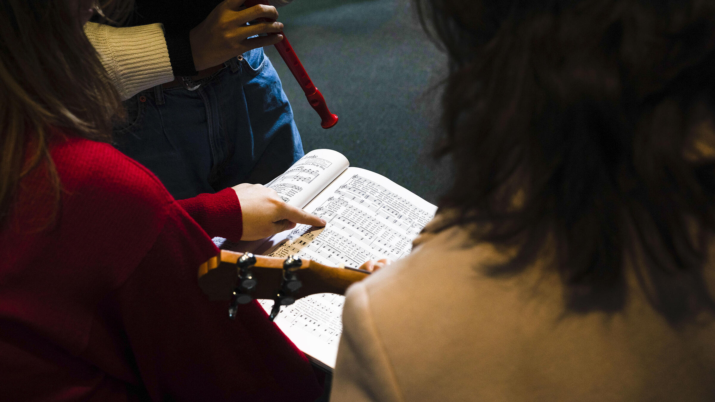 Menschen mit Instrumenten sitzen im Kreis und Finger zeigt auf gedruckte Noten in aufgeschlagenem DIN A4-Buch in Bibliothek