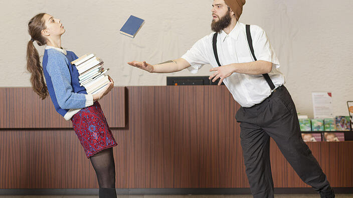 Frau mit Bücherturm auf Arm kippt nach hinten, während Mann sich zu ihr und fallendem Buch lehnt