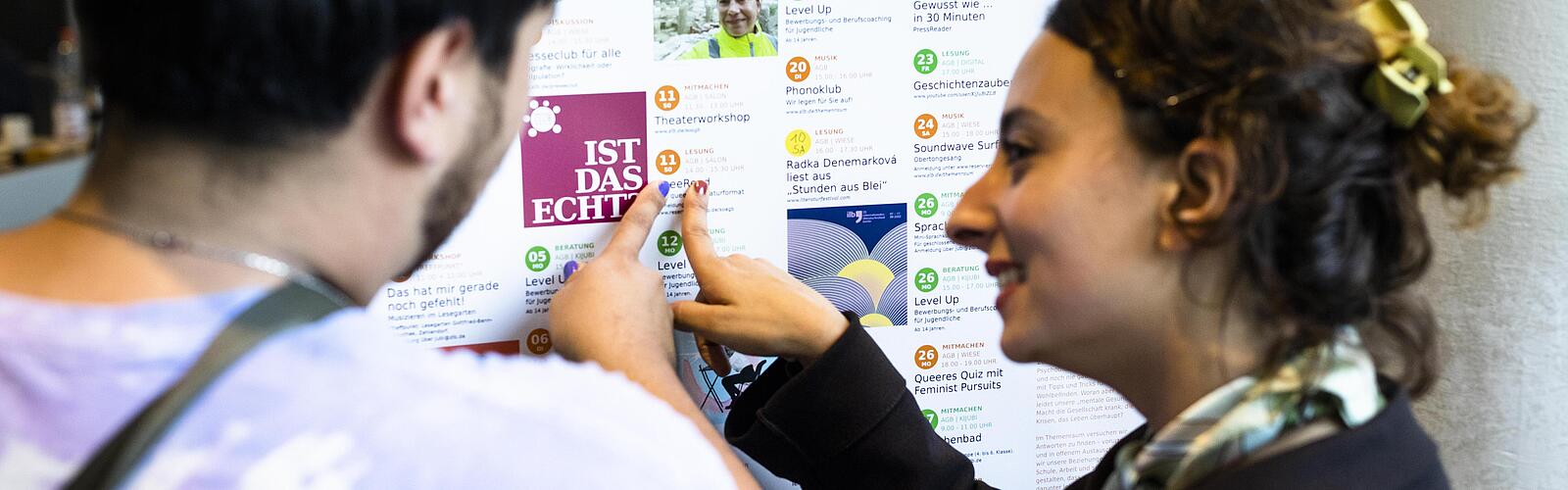 Zwei Menschen zeigen auf einen bunten Veranstaltungskalender mit kostenlosen Veranstaltungen in Berlin